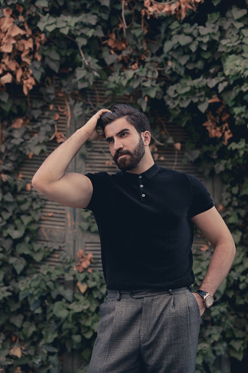 A fashionable young man poses confidently in front of an ivy-covered wall, showcasing modern style and confidence.