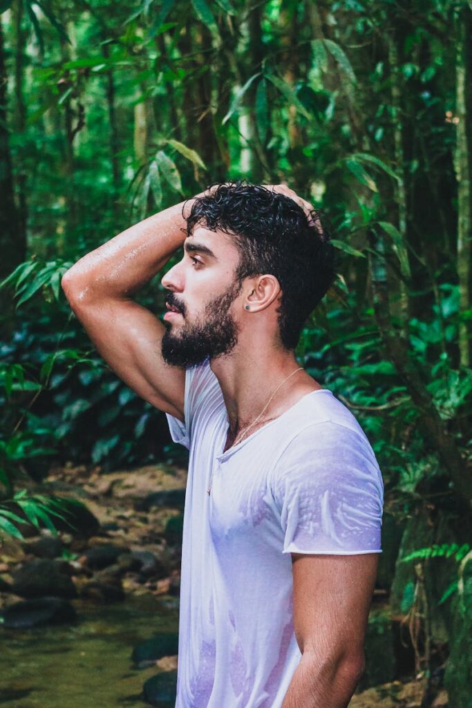 Profile of a handsome man in wet white shirt posing confidently in a lush jungle setting.