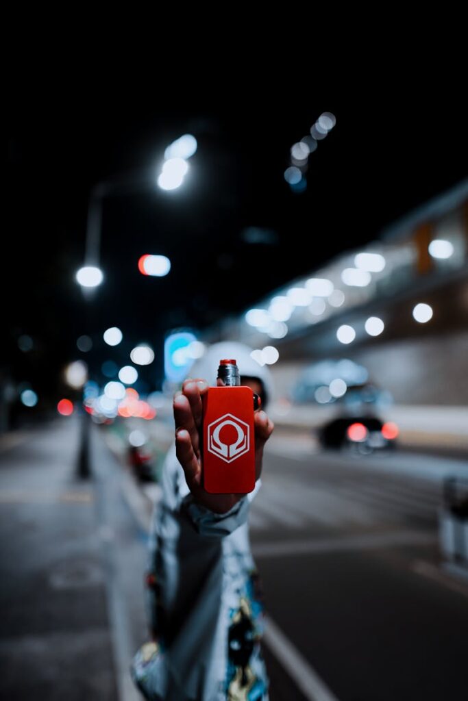 A red vape device held up against a blurred urban street scene with bright lights.